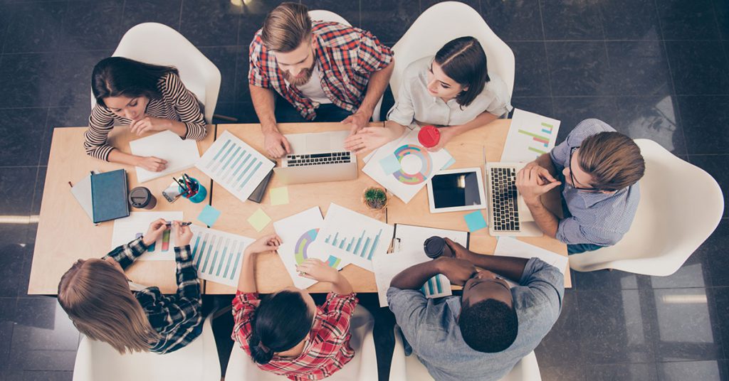 Overhead photo of seven people gathered about a table with spreadsheets and laptops